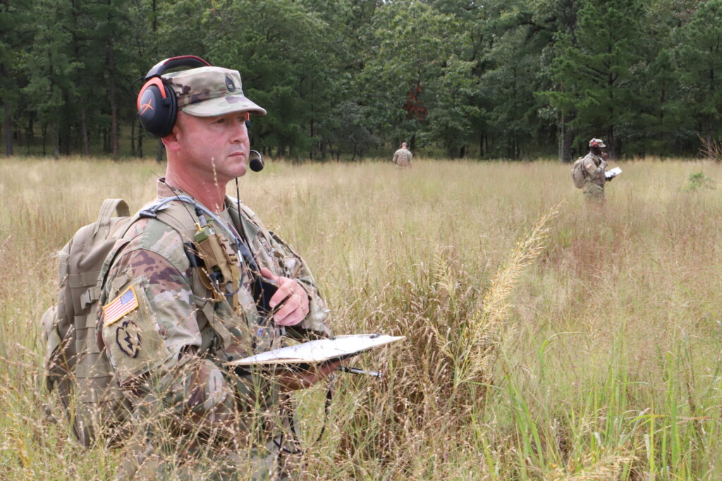 An American Soldier stands in a grass field wearing a radio headset and carrying a clip board. In the distance stand two other American Soldiers.